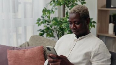 Afro-Amerikalı adam evdeki kanepede oturuyor ve akıllı telefon kullanıyor. Genç adam sosyal medyada sohbet ediyor. İnsanlar, teknoloji ve iletişim konsepti
