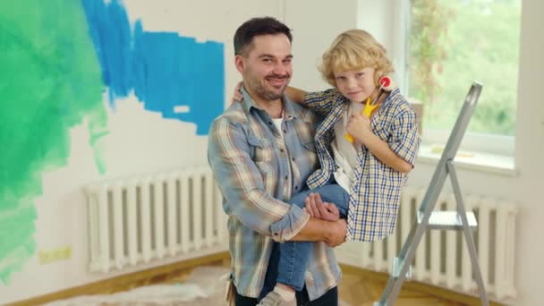 在新公寓里 父亲和儿子在一起的肖像 父亲牵着孩子 看着相机 而孩子牵着粉刷滚筒 人与流动的概念 — 图库视频影像