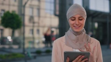 Happy arabian business woman wearing hijab going, working outdoors, swiping the social media. Young muslim lady working on tablet on the office centre background