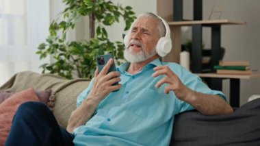 Mature caucasian man listening to music holding his phone while sitting on the sofa in living room and dancing while singing. Happy senior male singing and dancing while listening song