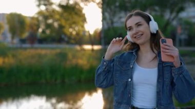 Kulaklıkla müzik dinleyen mutlu beyaz kadın, elinde akıllı telefon, parkta yürürken dans ediyor. Müzik dinlerken, kulaklık kullanarak dans eden güzel bir kadın.