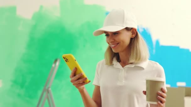 Женщина Работница Стоит Используя Смартфон После Покраски Стены Питья Кофе — стоковое видео