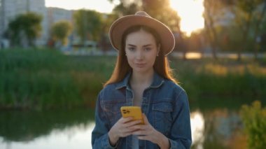 Akıllı telefon kullanan, kahverengi şapkalı, gölün arka planında kameraya bakan güzel kadının portresi. İnsanlar ve teknoloji kavramı