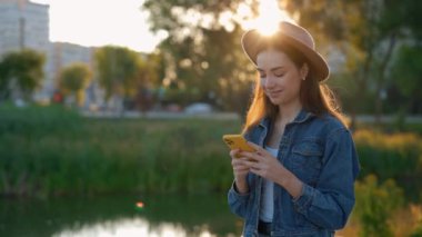 Güzel, gülümseyen beyaz bir kadın akıllı telefon kullanıyor, kahverengi şapka takıyor, göl kenarında yürüyor, mesajlaşıyor, uygulamaları kaydırıyor. İnsanlar ve teknoloji kavramı