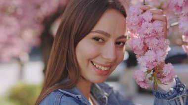 Portrait of beautiful young woman with sincere smile on face posing near blooming sakura tree. Attractive caucasian lady with long brown hair looking at camera