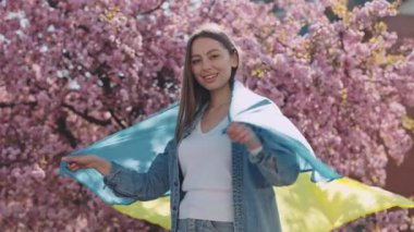 Patriotic young woman with Ukrainian flag on shoulders smiling and looking at camera while standing outside near sakura blooming trees. Proud of motherland, glory to Ukraine