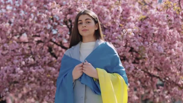 肩にウクライナのフラグを持つ桜の木の近くに立っている間にカメラを見て長い暗い髪を持つ若い女性の肖像画 祖国の誇りと支援の概念 — ストック動画