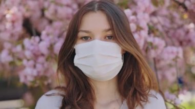 Portrait of woman in medical face mask looking at camera with background of blooming sakura tree. Pandemic prevention protective face mask during quarantine of coronavirus