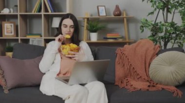 Charming pregnant woman sitting on cozy couch with bowl of crispy chips and watching movies on modern laptop. Harmful food during pregnancy. Expectant mother spending leisure time at home