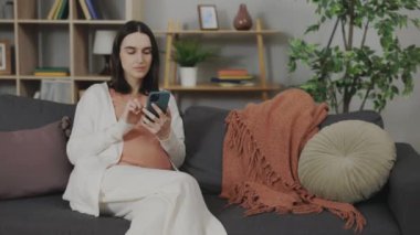 Young pregnant woman using smartphone for searching pregnancy information in internet. Future mom relaxing on comfy couch at living room with modern gadget in hands