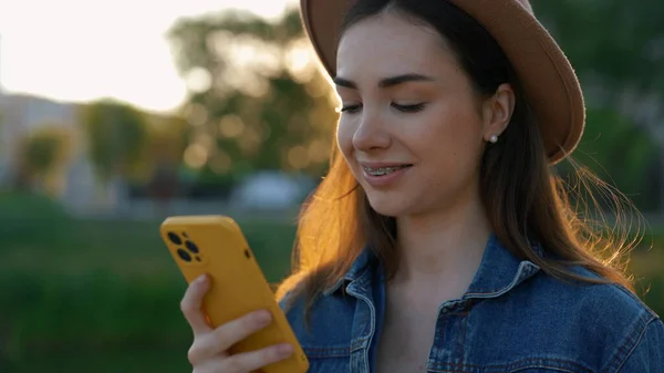 Beautiful caucasian woman with braces using smartphone, wearing brown hat, walking in the park texting, scrolling apps. People and technology concept