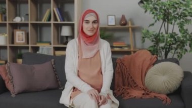 Attractive muslim women in hijab looking at camera and sincerely smiling while sitting on comfy sofa. Portrait of pregnant female relaxing and spending weekend at cozy home