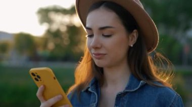 Beautiful caucasian woman with braces using smartphone, wearing brown hat, walking in the park texting, scrolling apps. People and technology concept