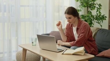 Young caucasian woman is studying online on laptop sitting on the sofa in her living room and makes notes. People, online education concept