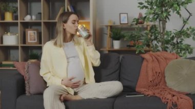 Caucasian pregnant woman in casual wear having glass of fresh milk while sitting on soft couch. Beautiful expectant lady enjoying healthy drink at cozy apartment.