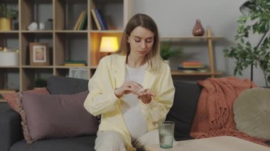 Young pregnant woman taking daily medicine, pills or complex of vitamins while sitting on comfy couch at home. Healthcare, pregnancy and treatment concept