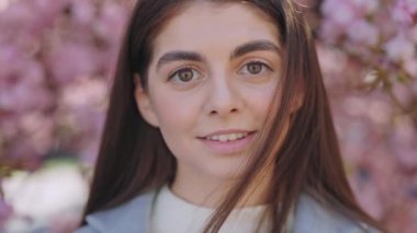 Close up portrait of attractive woman with brown hair and lush eyebrows smiling and looking at camera. Young female model with interesting appearance posing near sakura tree