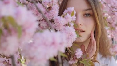 Sensual caucasian woman with brown hair covering half of face with branches of blooming sakura tree. Lady standing in the park. Seasonal flowering and femininity concept. Face portrait