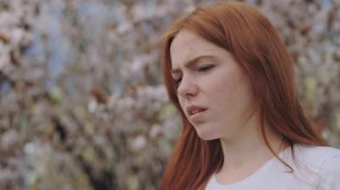 Close up of upset female having pollen allergy symptoms during spring outdoors. Frustrated caucasian lady rubbing her eyes while standing in front of blooming tree.