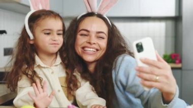Happy mother and daughter doing selfie with easter eggs, wearing bunny ears, have beauty and fun day together at home. Concept of childhood, technology, family weekend
