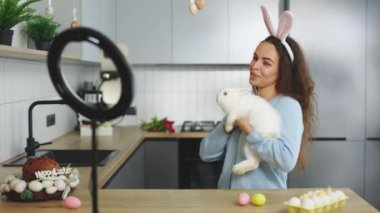 Happy woman conducts lesson of Easter symbol-rabbit, for video blog home. Female telling how to preparing for holidays. Display of smartphone recording video blog for easter vlog. Easter bunny
