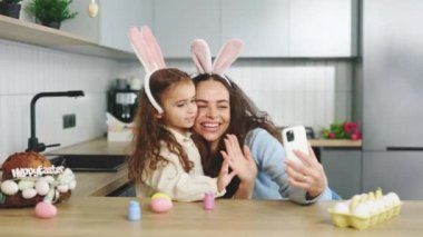 A cheerful family is preparing for the easter holiday using smartphone to video calling. Mom and daughter paint eggs, communicate with friends through video. Online talking. Happy easter