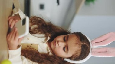 Vertical video. Beautiful little girl wearing bunny ears sitting in the kitchen table and using smartphone. Happy easter. Cute kid scrolling apps, watching videos on internet