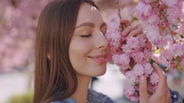 Sensual dark haired woman in casual wear touching branch with fresh spring flowers on sakura tree. Caucasian young lady standing outdoors, smiling and looking at camera.