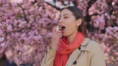 Charming young woman standing near blooming sakura tree and using medical spray to relief pain on throat outdoors. Concept of people and health problems