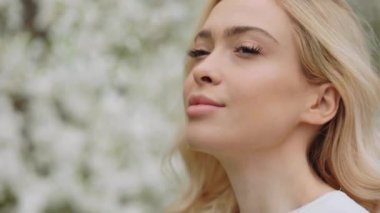 Close up of pretty young lady with wavy blond hair smelling young white flowers on apple trees at spring garden. Enjoyment from aroma. Nature and people concept.