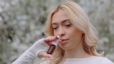 Young woman feeling unwell while being allergic to pollen. Close up of unhappy caucasian female treating rhinitis with nasal drops in blooming park