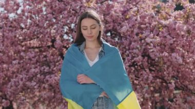 Proud Ukrainian woman with national flag on shoulders looking at camera with calm face while posing near sakura tree. Portrait. Girl lookina at camera. Power and strength of Ukrainian people