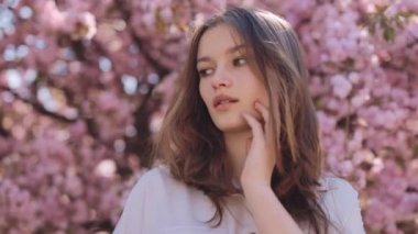 Charming portrait young woman with hand near chin standing in the blooming trees and looking at camera. Blur background of beautiful sakura with pink spring flowering