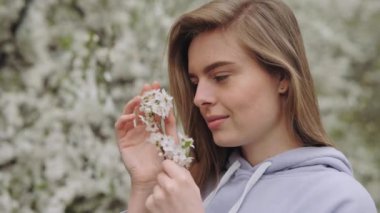 Tender young woman with cutted branch of blooming fruit tree in hands looking at camera standing in the park. Good-looking lady admiring beauty of nature during spring time