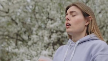 Unhealthy sick woman with allergy symptom blowing nose surrounded with seasonal flowering trees. Young caucasian lady having allergy symptoms outdoors