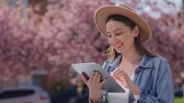 Caucasian attracrive woman walking outside on the blooming sakura trees background using digital tablet. Young lady browsing internet on the gagget. Socal media and people