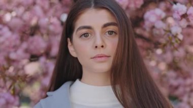 Portrait of charming young woman with pretty face looking at camera while standing over blooming sakura tree. Concept of people, femininity and springtime.