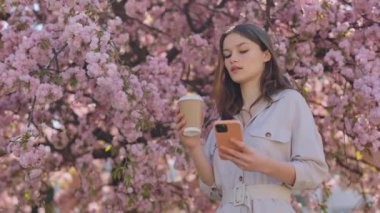 Beautiful young woman surfing internet on modern smartphone and enjoying fresh coffee on city street. Blur background of spring sakura flowering.