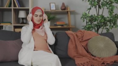 Pregnant arabian woman putting wireless headphones on belly while sitting on soft couch at cozy apartment. Positive expectant mother getting in touch with her child.
