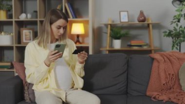 Smiling young pregnant woman using credit card with modern smartphone for shopping online at home. Preparation for maternity and technology concept.