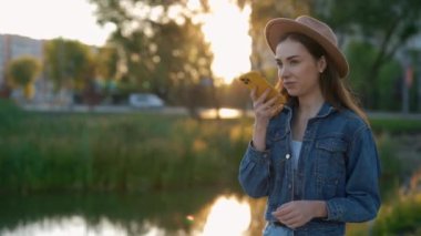 Beautiful young caucasian woman walking, wearing hat, using smartphone, recording voice message. Attractive lady on a walk, recording audio message to her friend