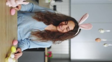Vertical video. Portrait of the happy woman wearing bunny ears headband standing in the decorated kitchen before painting the easter eggs. Holiday atmosphere. Smiling lady preparing for easter