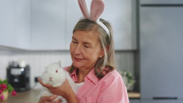 ふわふわのイースターウサギを持ち ウサギの耳をしている幸せな先輩女性の肖像画 笑顔の高齢者の女性は春休みの準備をしています イースターの準備 — ストック動画