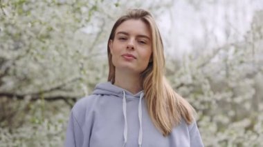 Portrait of shy smiling lady in casual blue hoodie standing among blossoming trees in park. Young woman feeling happiness while relaxing from city bustle.