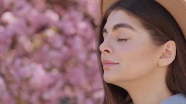 Side view of caucasian young woman wearing trendy beige hat posing outdoors with blur background of blooming sakura tree. Pleasant dark haired female enjoying smell of seasonal flowering.