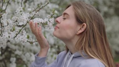 Side view of pretty lady sniffing and touching beautiful white flowers on branches. Young gorgeous female smiling and looking at camera while enjoying blossoming tree aroma in spring park.