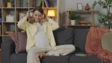 Relaxed woman in expectation sitting on couch listening music in wireless headphones. Young pregnant lady sharing favorite song with her unborn baby in big belly. Connection between mother and child