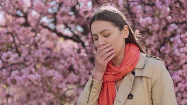 公園の外に立っている間 喉の痛みに苦しむスカーフを身に着けている暗い髪の原因アジアの女性 春の開花時に季節のアレルギーを持つ若い女性 — ストック動画