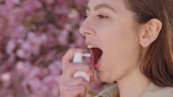 医学スプレーを使用して喉の痛みを気にUnwell若い女性の側面図 春の桜の木の近くに立っている白人ブルネットとアレルギーに苦しんで — ストック動画