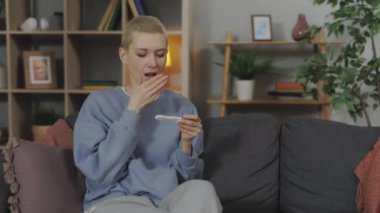 Surprised caucasian woman checking pregnancy test while sitting on couch. Beautiful lady with short blond hair feeling happiness during long-awaited pregnancy.